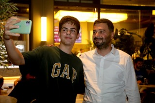 Mevlüt Öztekin gece bir kafede, telefonuyla özçekim yapan genç bir hemşehrimizle poz veriyor.