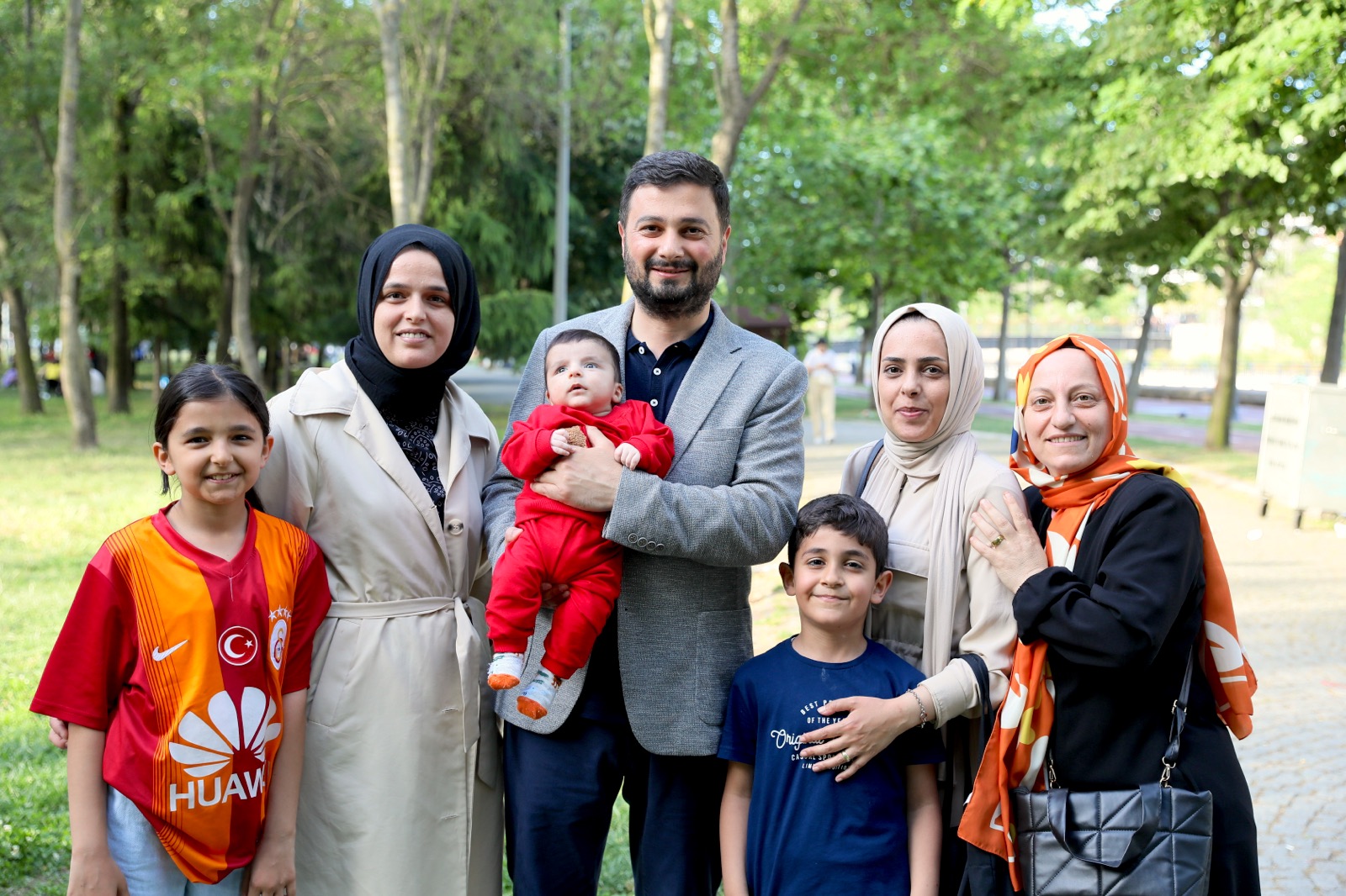 Mevlüt Öztekin parkta kırmızı tulumlu bir bebeği kucağında tutuyor, çevresinde üç kadın ve iki çocuk var.