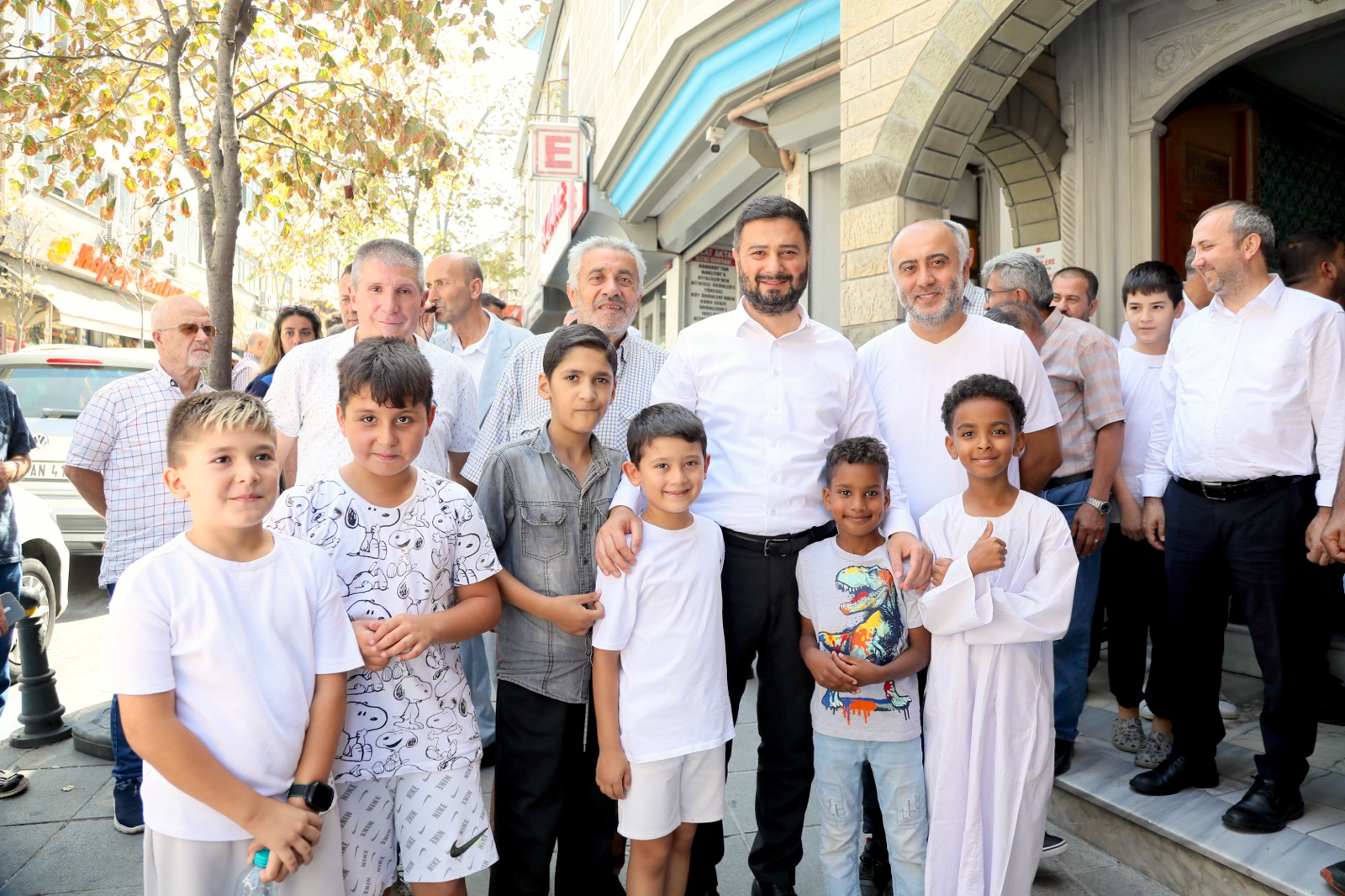 Mevlüt Öztekin taş kemerli bir girişin önünde çocuklar ve yetişkinlerden oluşan kalabalık bir grupla poz veriyor.