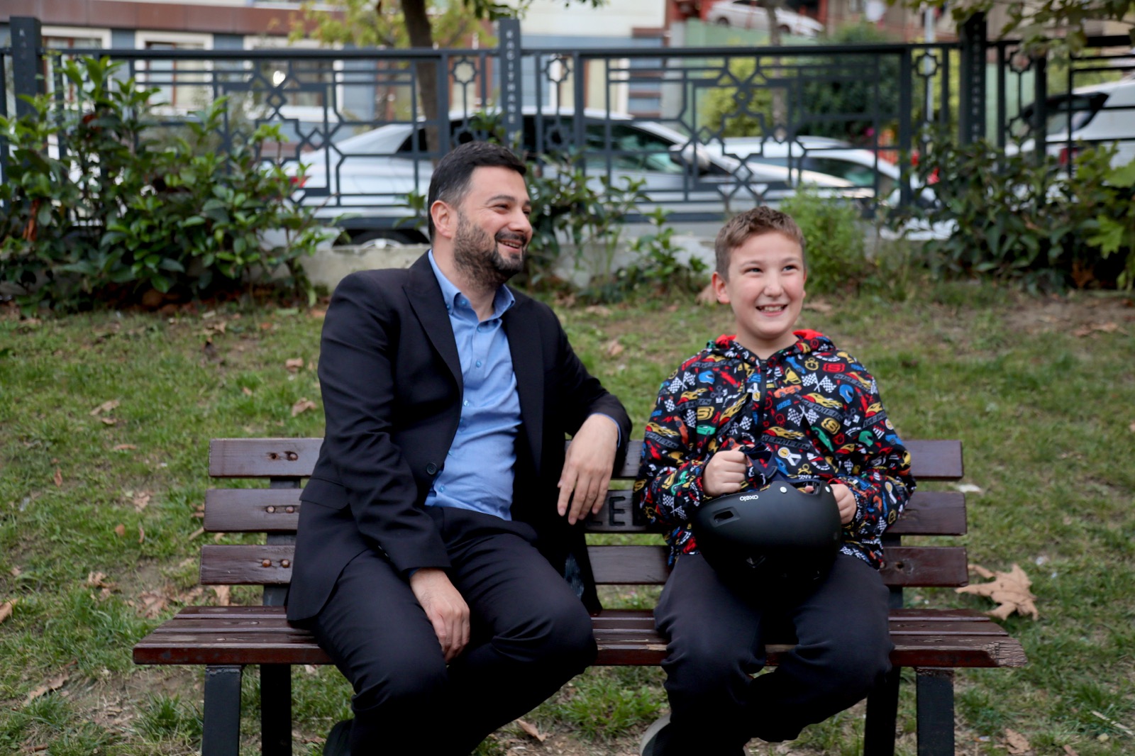 Mevlüt Öztekin parkta bir bankta, elinde bisiklet kaskı tutan gülen bir çocukla oturuyor.