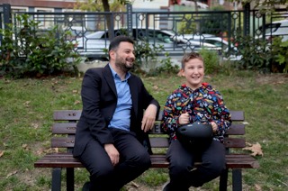 Mevlüt Öztekin parkta bir bankta, elinde bisiklet kaskı tutan gülen bir çocukla oturuyor.