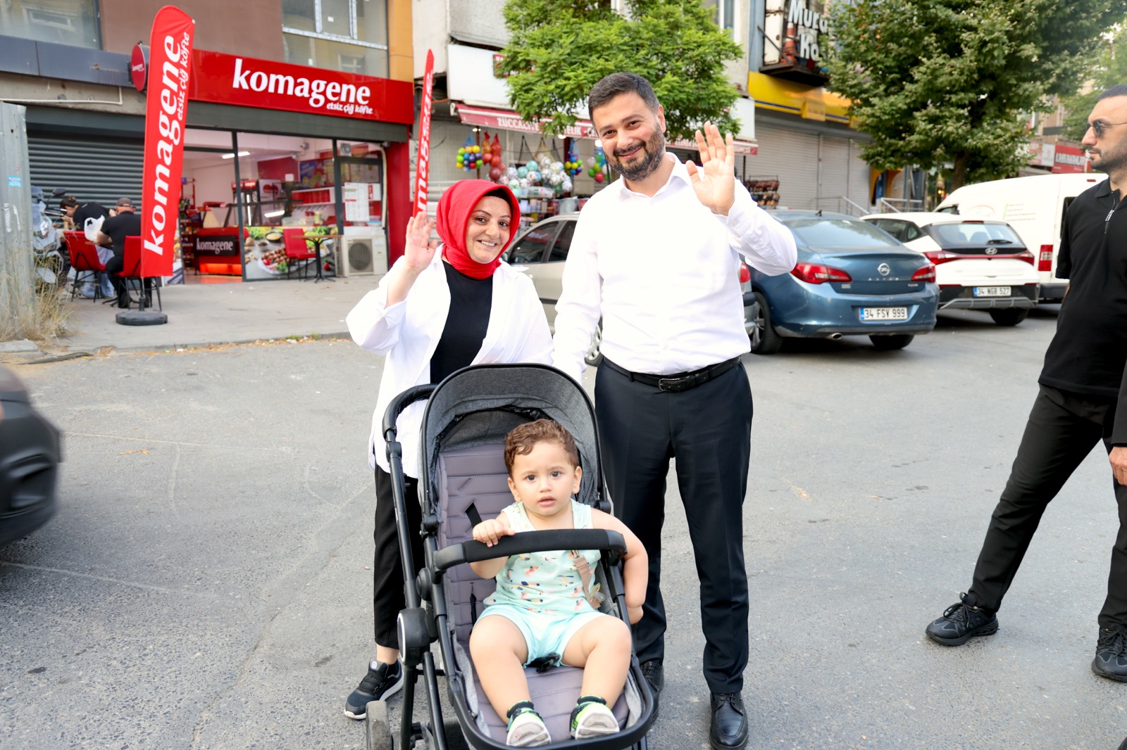 Mevlüt Öztekin sokakta bebek arabasında bir çocukla duran bir kadınla birlikte el sallıyor.