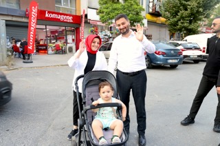Mevlüt Öztekin sokakta bebek arabasında bir çocukla duran bir kadınla birlikte el sallıyor.