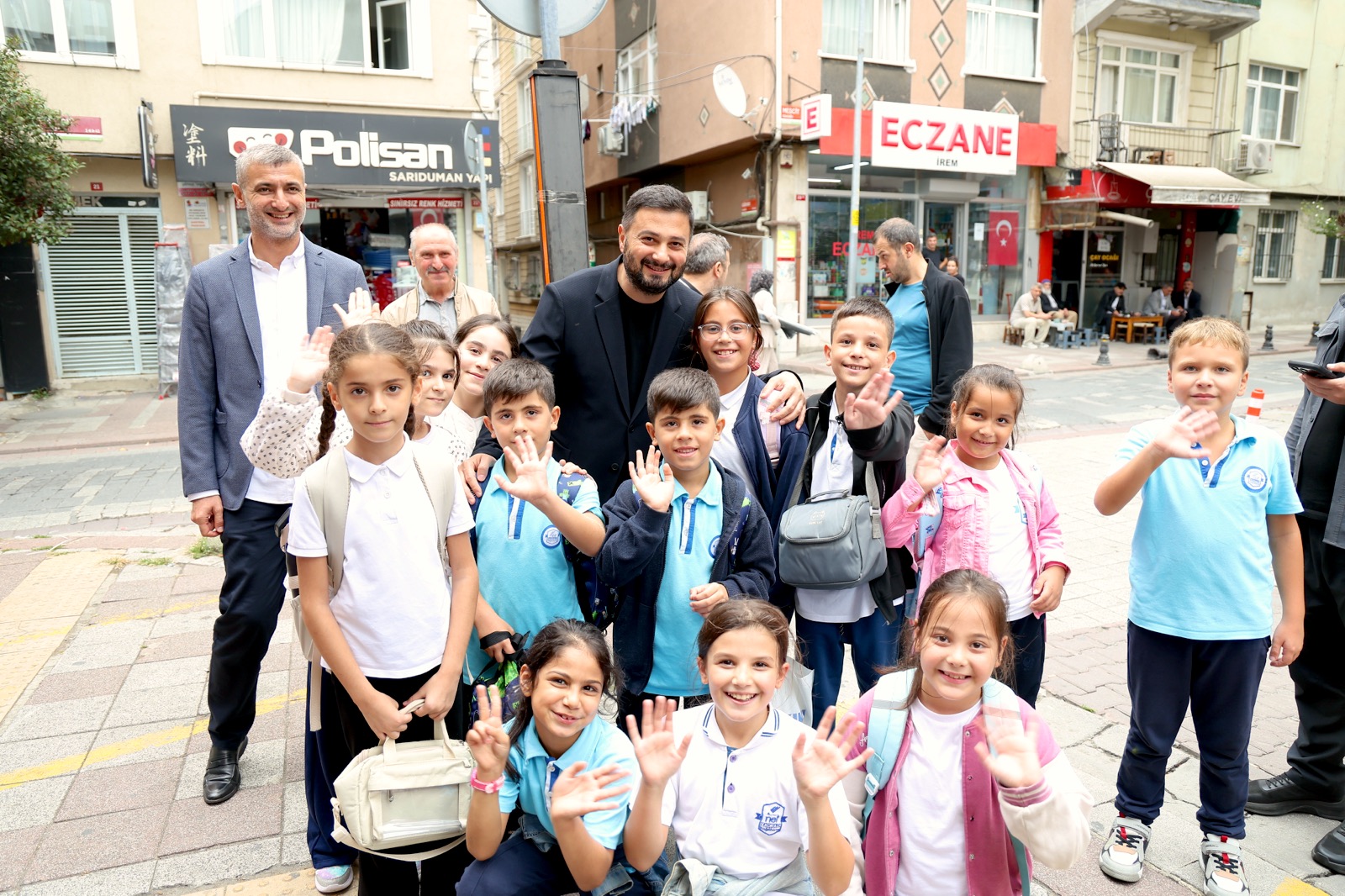 Mevlüt Öztekin bir eczanenin önünde okul formalı, el sallayan çocukların arasında gülümsüyor.