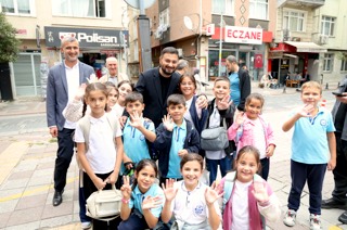 Mevlüt Öztekin bir eczanenin önünde okul formalı, el sallayan çocukların arasında gülümsüyor.