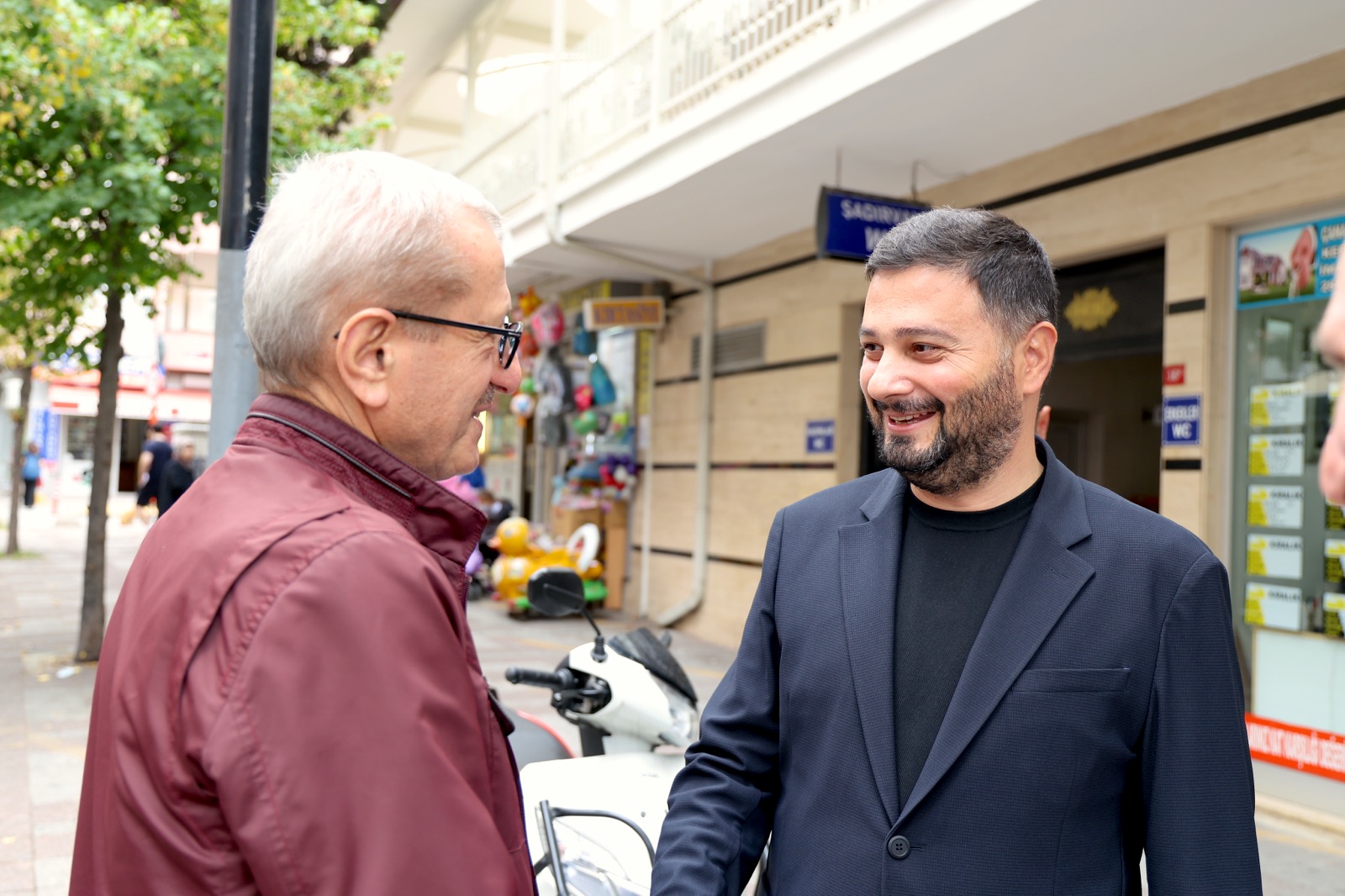 Mevlüt Öztekin kaldırımda gözlüklü yaşlı bir hemşehrimizle gülümseyerek sohbet ediyor.