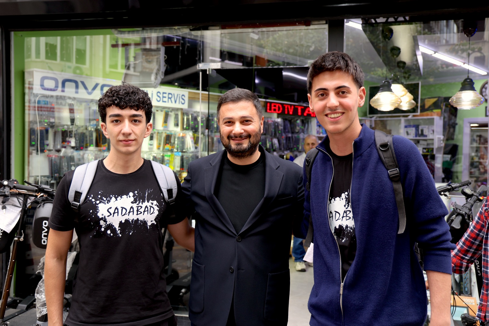 Mevlüt Öztekin bir elektronik dükkânının vitrini önünde sırt çantalı iki gençle poz veriyor.