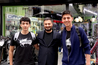 Mevlüt Öztekin bir elektronik dükkânının vitrini önünde sırt çantalı iki gençle poz veriyor.