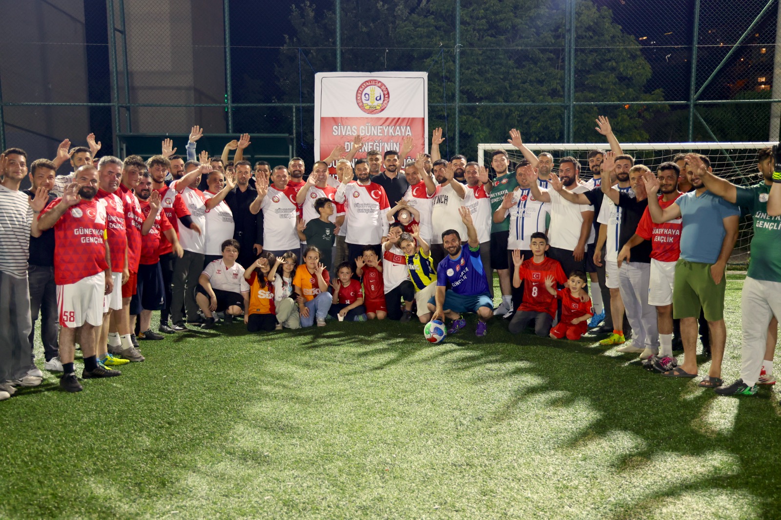 Mevlüt Öztekin gece bir halı sahada formalı futbolcular ve çocuklarla el sallayarak toplu fotoğrafta.
