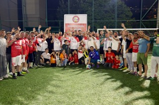 Mevlüt Öztekin gece bir halı sahada formalı futbolcular ve çocuklarla el sallayarak toplu fotoğrafta.