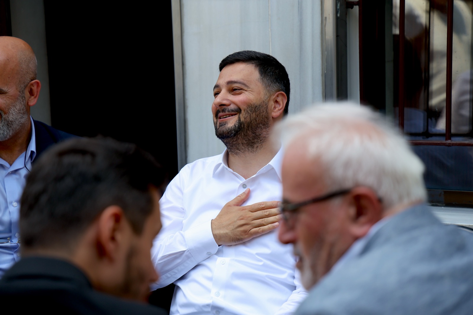 Mevlüt Öztekin oturduğu yerde elini göğsüne koymuş gülümsüyor, önde sohbet eden hemşehrilerimiz bulanık.