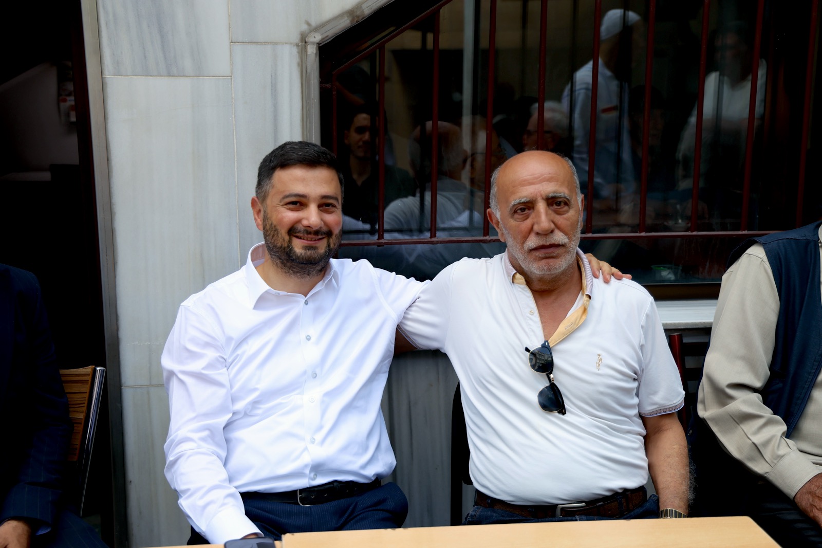 Mevlüt Öztekin mermer duvarlı bir avluda oturmuş, kolunu yaşlı bir hemşehrimizin omzuna atmış.