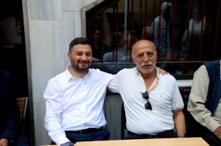 Mevlüt Öztekin mermer duvarlı bir avluda oturmuş, kolunu yaşlı bir hemşehrimizin omzuna atmış.