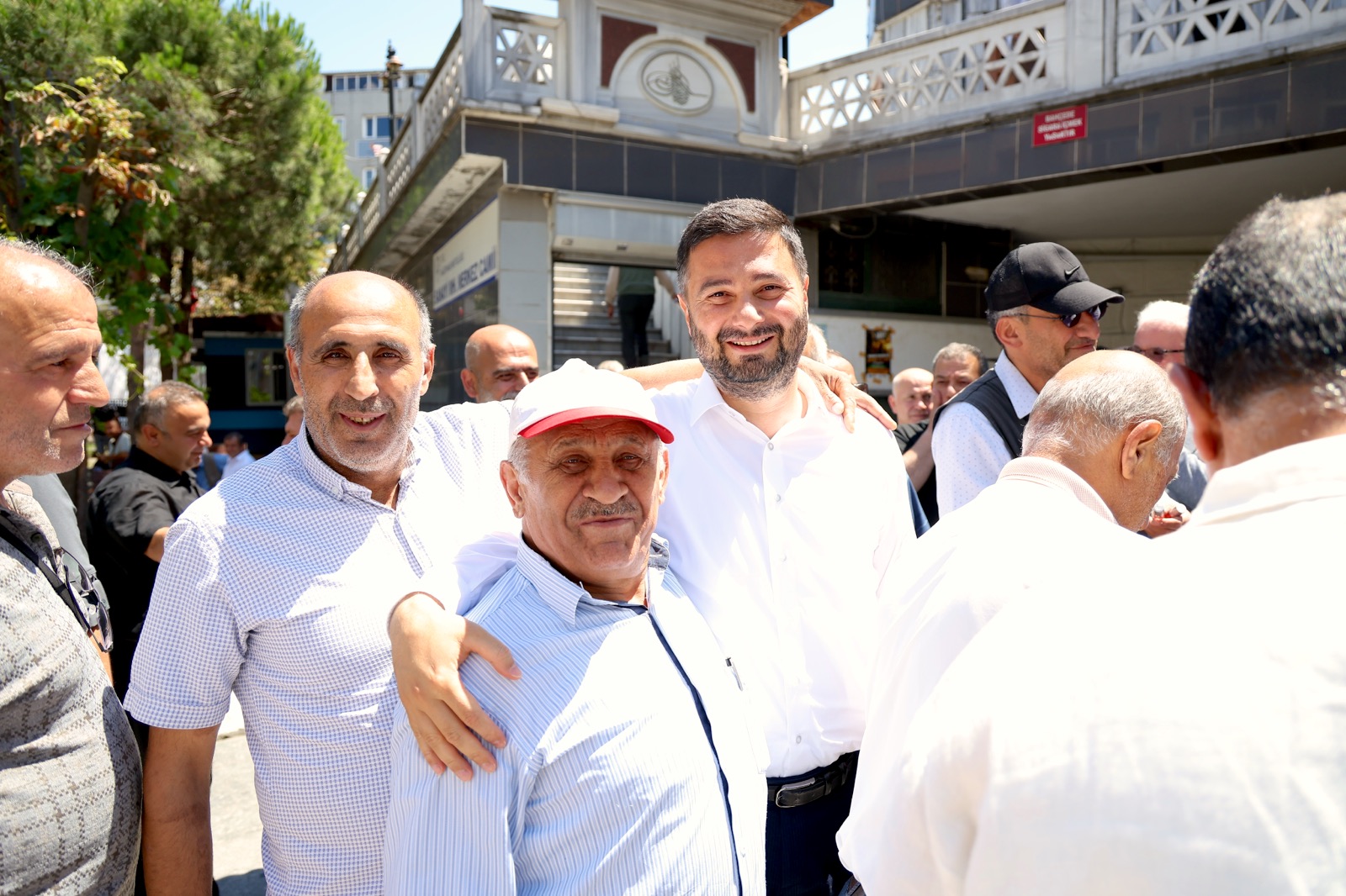 Mevlüt Öztekin bir caminin önündeki kalabalıkta şapkalı yaşlı bir hemşehrimize sarılmış gülümsüyor.