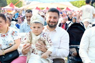 Mevlüt Öztekin seyirciler arasında oturmuş, kucağında sünnet kıyafetli ve oyuncak kılıçlı bir çocuk tutuyor.