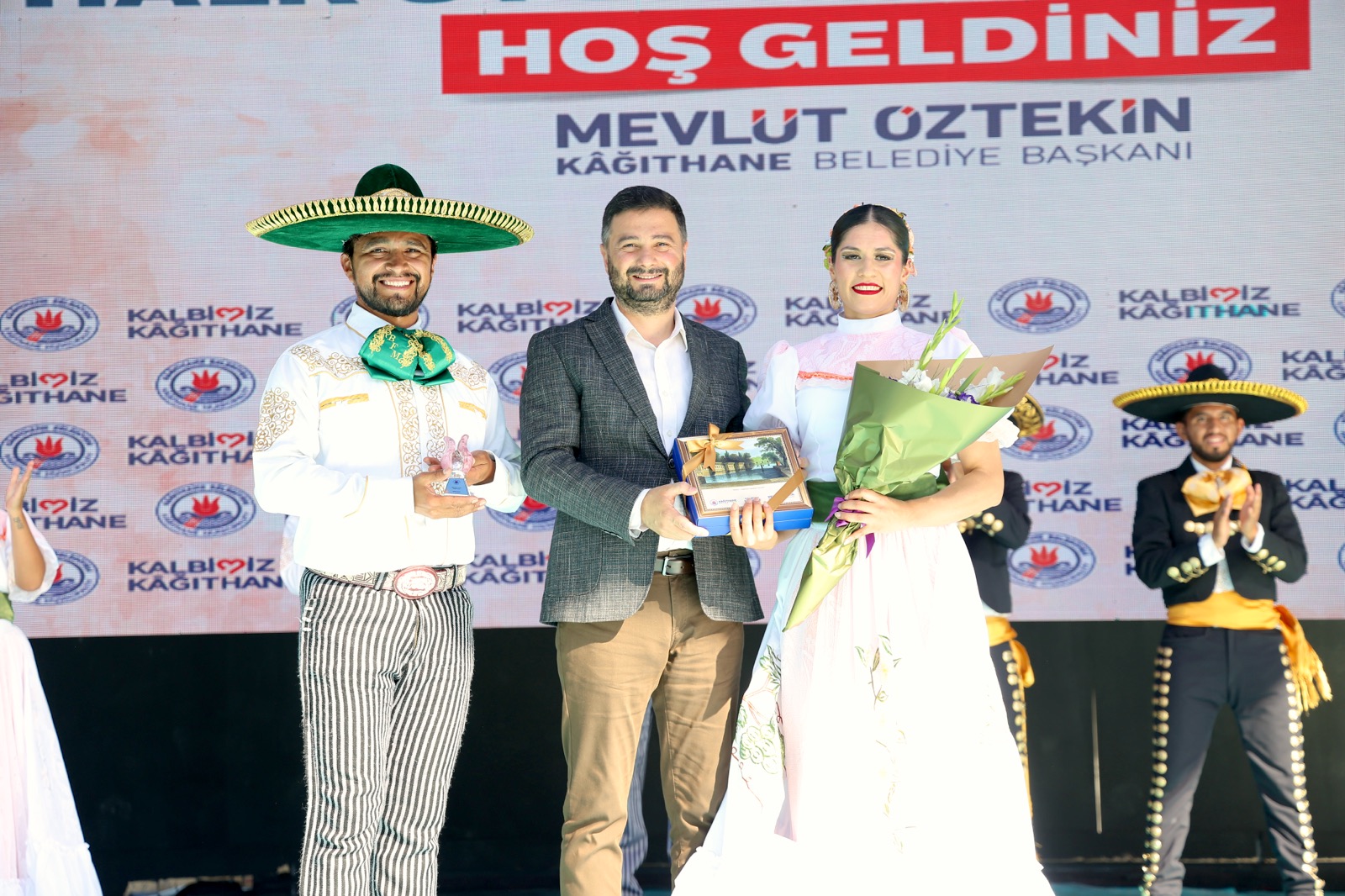 Mevlüt Öztekin sahnede geniş şapkalı ve uzun etekli iki halk dansçısıyla, elinde bir hediye kutusu tutuyor.