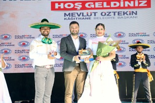 Mevlüt Öztekin sahnede geniş şapkalı ve uzun etekli iki halk dansçısıyla, elinde bir hediye kutusu tutuyor.