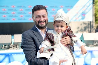 Mevlüt Öztekin sünnet kıyafeti giymiş, elinde oyuncak kılıç tutan küçük bir çocuğu kucağında taşıyor.