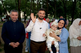 Mevlüt Öztekin parkta el sallıyor, yanında sünnet kıyafetli bir bebeği kucağında tutan bir kadın gülümsüyor.