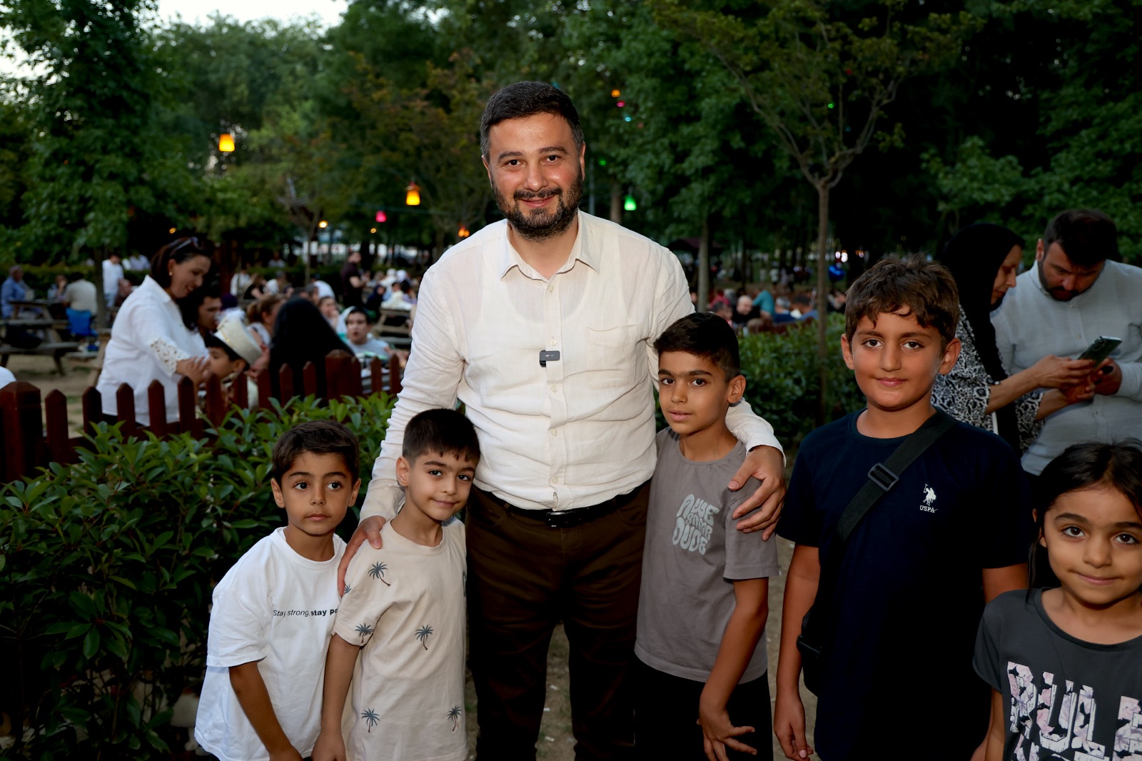Mevlüt Öztekin akşam ışıklarıyla süslenmiş bir parkta beş çocukla birlikte poz veriyor.