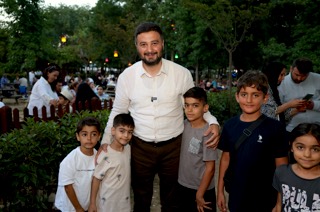 Mevlüt Öztekin akşam ışıklarıyla süslenmiş bir parkta beş çocukla birlikte poz veriyor.