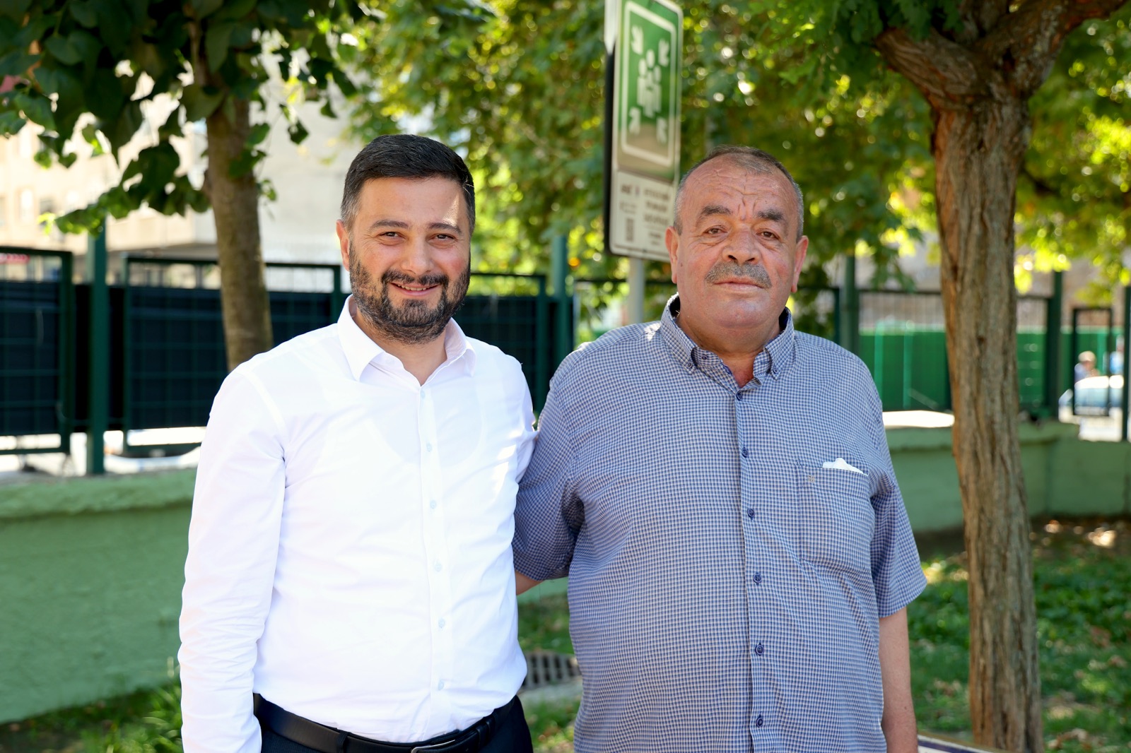 Mevlüt Öztekin ağaçların altında kareli gömlekli yaşlı bir hemşehrimizle yan yana poz veriyor.