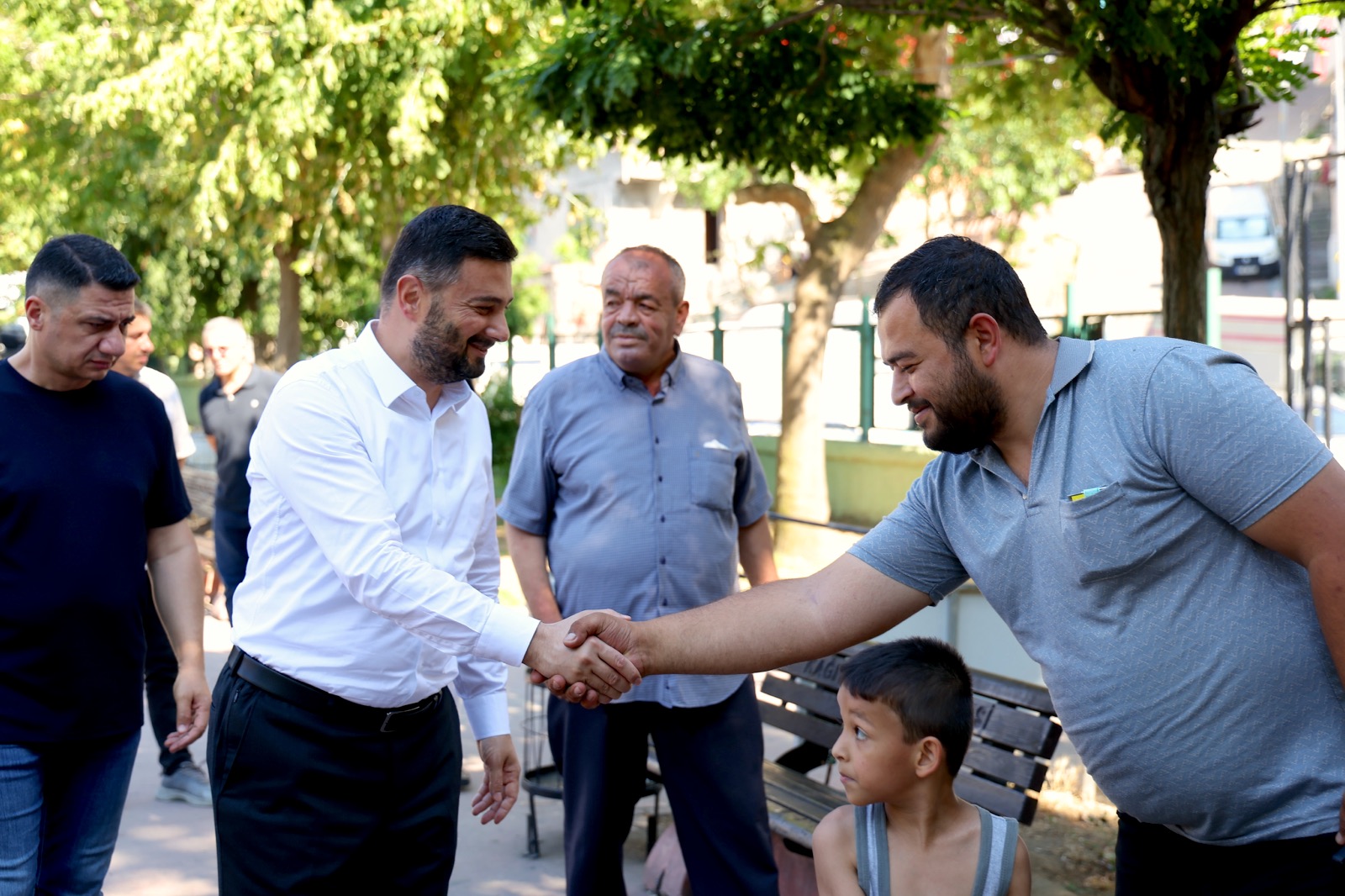 Mevlüt Öztekin parkta bir hemşehrimizle tokalaşıyor, bankta oturan bir çocuk onlara bakıyor.
