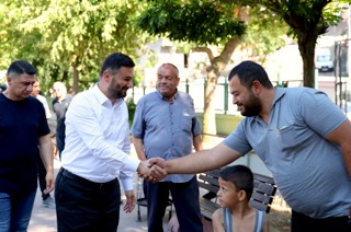 Mevlüt Öztekin parkta bir hemşehrimizle tokalaşıyor, bankta oturan bir çocuk onlara bakıyor.