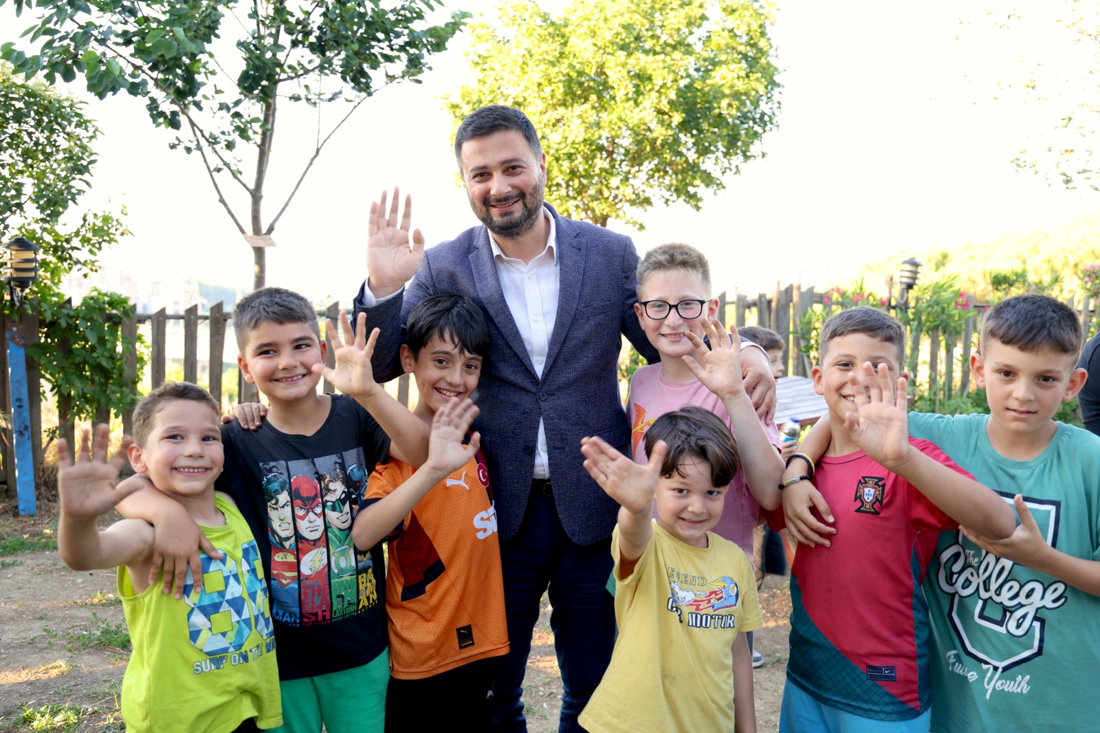 Mevlüt Öztekin bir bahçede el sallayarak gülen yedi çocuğun ortasında, o da el sallıyor.
