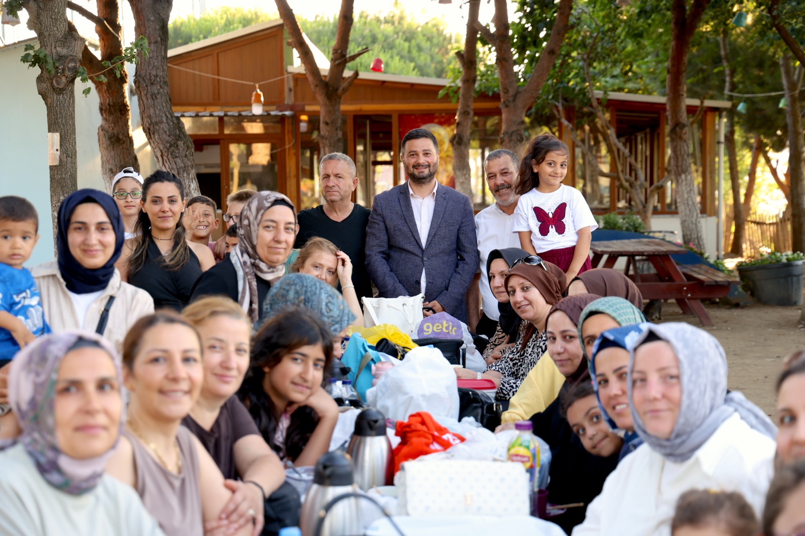 Mevlüt Öztekin piknik masaları çevresinde oturan kadınların ve çocukların arkasında ayakta duruyor.