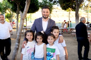 Mevlüt Öztekin piknik masalarının olduğu bir alanda kollarını dört çocuğun omzuna dolamış gülümsüyor.