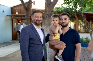 Mevlüt Öztekin ağaç altında, kucağında küçük bir çocuk taşıyan genç bir adamla yan yana poz veriyor.