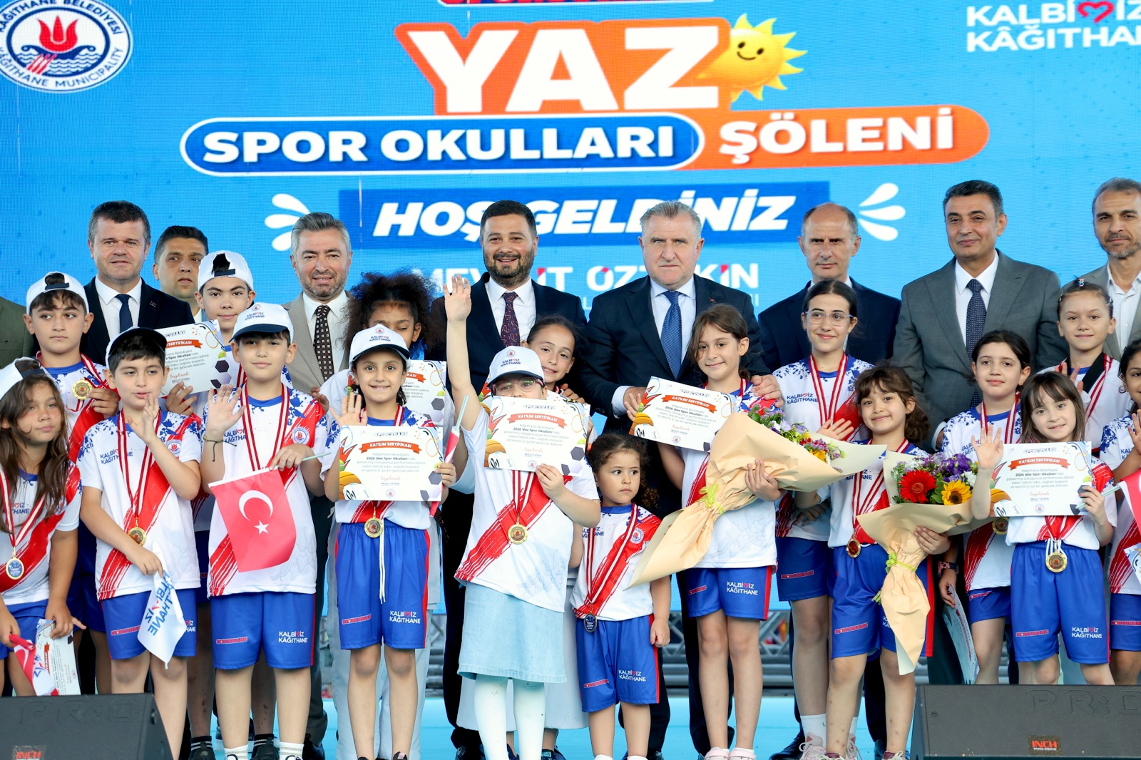 Mevlüt Öztekin sahnede madalyalı ve sertifikalı sporcu çocuklar ve protokol üyeleriyle toplu fotoğrafta.