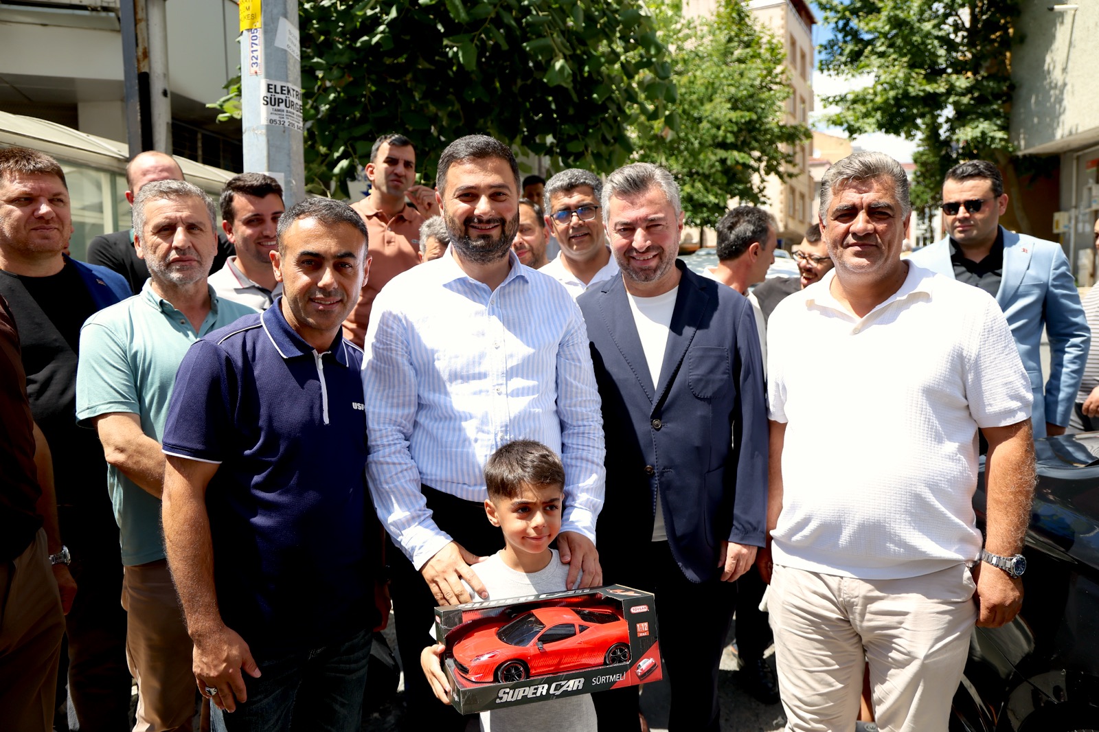 Mevlüt Öztekin sokakta kalabalığın ortasında, kutulu oyuncak araba tutan bir çocuğun omuzlarına ellerini koymuş.