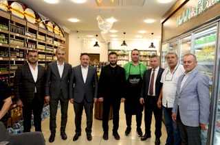 Mevlüt Öztekin raflarla dolu bir şarküteride önlüklü bir çalışan ve misafirlerle sıra hâlinde poz veriyor.